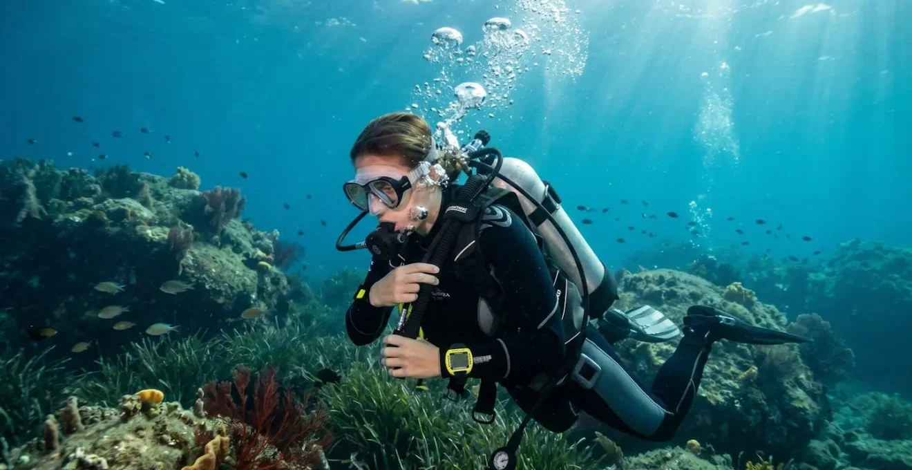Un plongeur en combinaison néoprène ajuste son gilet stabilisateur en pleine eau, vu de profil, avec des bulles d'air montant vers la surface et un fond récifal flou en arrière-plan