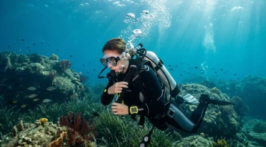 Un plongeur en combinaison néoprène ajuste son gilet stabilisateur en pleine eau, vu de profil, avec des bulles d'air montant vers la surface et un fond récifal flou en arrière-plan